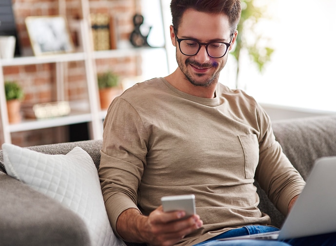Hombre sentado en el sofá mirando su teléfono mientras tiene una portátil en el regazo